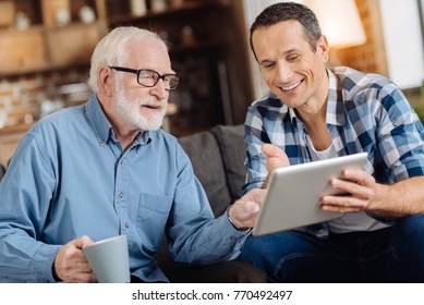 Help needed. Pleasant senior man sitting on the sofa next to his son and pointing at the tablet, asking his son to explain how to use the tablet - Powered by Shutterstock