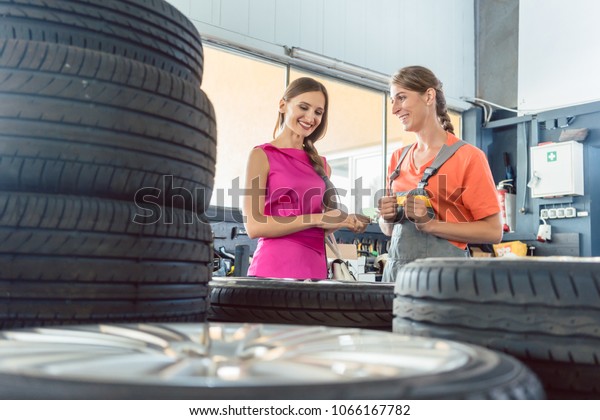 Helpful experienced female auto mechanic\
checking the identification number of a tire for a customer in an\
automobile repair shop with various tires for\
sale