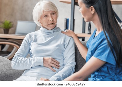 Helpful female nurse wearing uniform listening elderly patient at home, while old grandmother complaining about belly stomach ache pain. Treatment and medicine, drugs medications prescription - Powered by Shutterstock