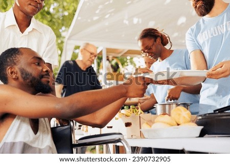 Helpful guy volunteer aids and provides free food to a poor needy african american man in wheelchair. Volunteers at an outdoor food bank provide hunger assistance to the  underprivileged.