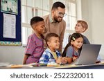 Helpful male professor talking to students while using computer in primary classroom. Male teacher supervising a group of elementary children working on laptop at school. 