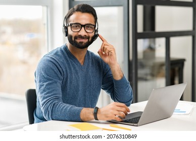 Helpful And Smiling Indian Male Employee Wearing Headset And Eyeglasses Making Online Consultation, Holding Video Call, Man In Touch Looks At Camera And Touching Button On Headphones, Accepts A Call