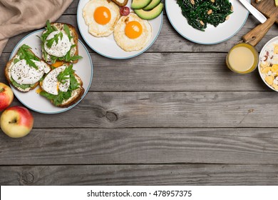 Helpful and tasty breakfast from different of dishes - fried egg, poached eggs, avocado, apple, spinach salad, muesli and orange juice on wooden table with copy space, top view