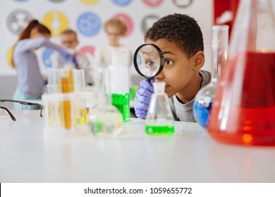 Helpful tool. Upbeat teenage boy looking at a chemical flask through a magnifying glass while examining the reaction going on in the flask
