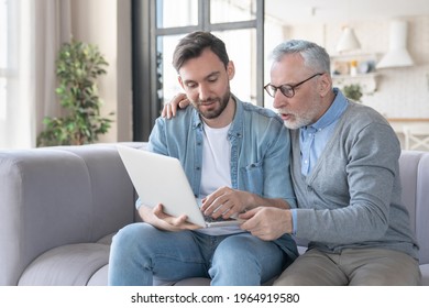 Helpful young adult son helping aiding his old senior elderly father in using laptop for watching movies, communicating online. Son teaching father how to use laptop. Happy father`s day!