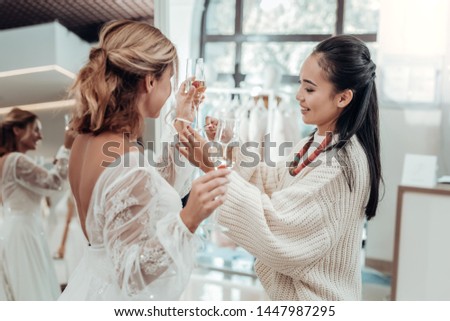 Helping a friend. Beautiful bride holding glasses of champagne while her bridesmaid helping her trying a wedding dress.