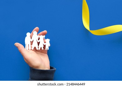 Helping hand holding Ukrainian family, white paper silhouette. Flat lay, top view. Copy-space, text place on blue background with yellow ribbon. Creative concept paper art. Volunteer help, refugees.