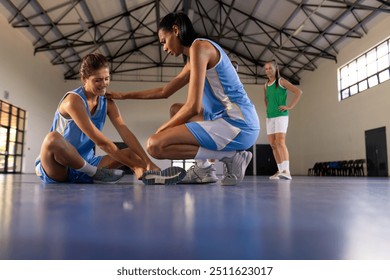 Helping injured teammate, female basketball players in gym during practice session. Teamwork, support, coaching, injury, fitness, recovery - Powered by Shutterstock