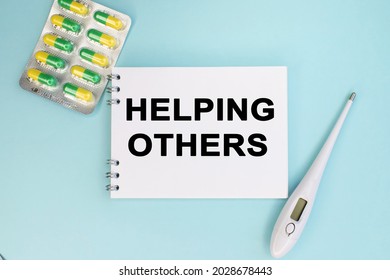 Helping Others Is Written On A Notebook That Lies On A Blue Background Near A Thermometer And Tablets In A Blister. Medical Concept