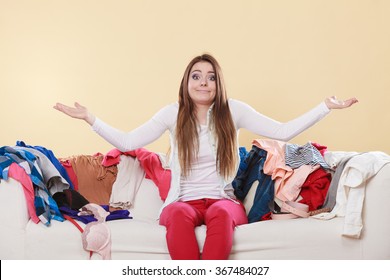 Helpless Woman Sitting On Sofa Couch In Messy Living Room Shrugging. Young Girl Surrounded By Many Stack Of Clothes. Disorder And Mess At Home.