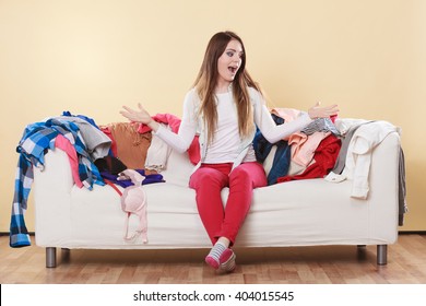 Helpless Woman Sitting On Sofa Couch In Messy Living Room Shrugging. Young Girl Surrounded By Many Stack Of Clothes. Disorder And Mess At Home.