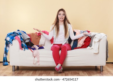 Helpless Woman Sitting On Sofa Couch In Messy Living Room Shrugging. Young Girl Surrounded By Many Stack Of Clothes. Disorder And Mess At Home.