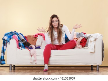 Helpless Woman Sitting On Sofa Couch In Messy Living Room Shrugging. Young Girl Surrounded By Many Stack Of Clothes. Disorder And Mess At Home.