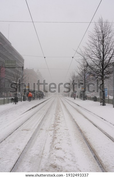 Helsinki, Finland 03 09 2021, Snowstorm and\
snow-covered street,  lonely pedestrian, Persons crossing the\
street blurred snowy, focus on foreground snowflakes, tram-train, a\
man with face mask\
covid19