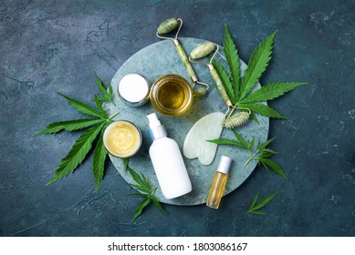 Hemp leaves, oil, cosmetic products, face cream, body butter, face roller and gua sha massager on dark background. Top view, copy space. Natural skin and self care concept. Flat lay. Banner