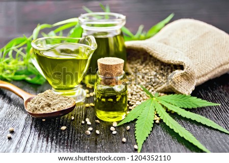 Hemp oil in two glass jars and sauceboat with grain in the bag, leaves and stalks of cannabis, a spoon with flour on the background of wooden boards