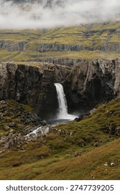 Hengifoss waterfall cascading down dramatic layered basalt cliffs with red volcanic strata in East Iceland, powerful stream falling into a rugged remote landscape under cloudy sky.