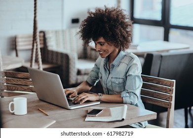 Her job is her life. Beautiful young African woman using computer and smiling while sitting at her working place in cafe - Shutterstock ID 617982719