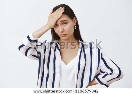 Her sister is like pain in ass. Tired and gloomy cute woman in striped blouse, holding palm on forehead, feeling under pressure, being exhausted and gazing upset at camera over gray background
