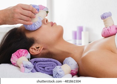 Herbal ball face massage in ayurveda spa. Female massagist with young woman in wellness center. Healthcare therapy to beautiful indian girl in beauty parlor, side view of face with eyes closed