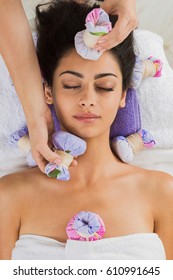 Herbal ball massage in ayurvedic spa. Female massagist with young woman in wellness center. Healthcare therapy to beautiful indian girl in beauty parlor, top view of face with eyes closed