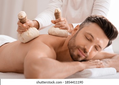 Herbal massage concept. Unrecognizable spa attendant making body massage for handsome man with herbal compress balls, luxury spa interior. Man enjoying herbal treatment at massage salon