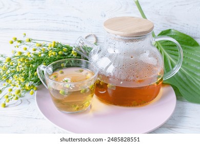 Herbal medicinal tea with chamomile flowers in a glass teapot, a bouquet of fresh flowers, on a wooden light background.