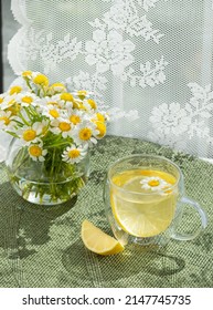 Herbal tea in a glass teacup with chamomile and lemon slice on green placemat. Chamomile lemon tea. Daisy fresh wild flowers in glass jar. White tulle background in sunlight, patterned shadows - Shutterstock ID 2147745735