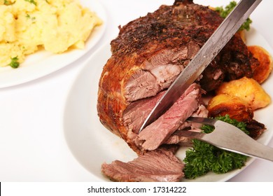A herbed, boneless leg of lamb being carved, with parsley and roast potatoes on the plate and mashed parsley potatoes in the background