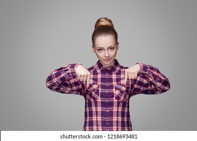 Here and right now. serious angry blonde girl in red, pink checkered shirt, collected bun hairstyle standing and looking at camera and pointing down. indoor studio shot. isolated on gray background