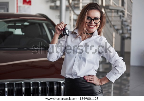 Here\'s your keys. Woman in eyewear and white\
shirt stands near the red\
automobile.