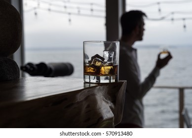 Hero Shot Of Whiskey On The Rocks With Silhouetted Man In Background Holding Glass.