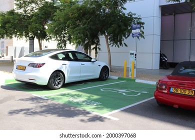 HERZLIYA, ISRAEL - JUNE 14, 2021: Parking Spot In Green, Part Of An EV Electric Vehicle Charging Station In Herzliya Pituach Industrial Zone. Charging Icon On The Parking Spot. Left: White Tesla Car.