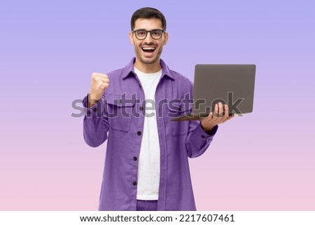 He's a winner! Happy young man in purple shirt looking at laptop screen with victory expression, isolated on purple background
