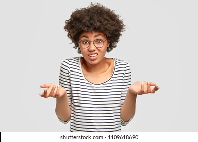 Hesitant African American Female Models In Studio, Has Puzzled Expression, Shruggs Shoulders With Uncertainty, Frowns Face. Doubtful Woman Costumer Can`t Make Choice, Isolated On White Wall.