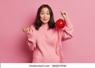 Hesitant Brunette Asian Woman Shrugs Shoulders And Cannot Decide How To Decorate Christmas Tree Hods Big Red Glass Bauble Wears Sweater Poses Against Pink Background. New Year Decor Concept.