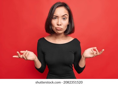 Hesitant Confused Woman Spreads Palms Looks Indignant Feels Unaware Wears Black Dress Raises Eyebrows Poses Against Red Background Shrugs Shoulders Doesnt Know What To Do. People Uncertainty