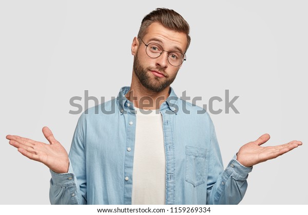 困惑した恥ずかしくない人間の肩をすくめ 決め手がなく 剛毛 トレンディな髪型 白い背景に青いスタイリッシュなシャツを着ている 室内で無知な男性のポーズ の写真素材 今すぐ編集