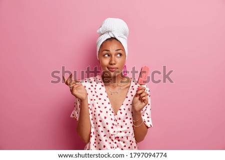 Hesitant thoughtful dark skinned woman with towel on head, enjoys eating tasty cold strawberry ice cream, has dreamy expression, wears pajama, poses against pink background. Domestic lifestyle