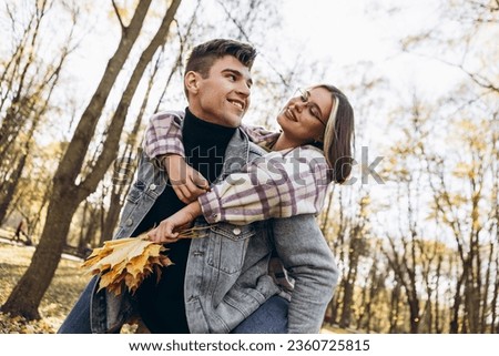 Heterosexual caucasian young loving couple, man carrying on back woman in sunny weather, hugging smiling kissing laughing spending time together. Autumn, fall season, orange yellow red maple leaves
