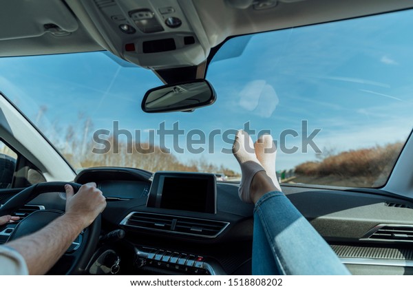 Heterosexual couple in a luxury\
car, making a road trip, the man has his hands on the steering\
wheel and the woman has her feet with some socks on top of the\
dashboard.