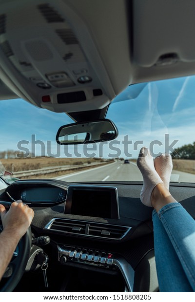 Heterosexual couple in a luxury\
car, making a road trip, the man has his hands on the steering\
wheel and the woman has her feet with some socks on top of the\
dashboard.