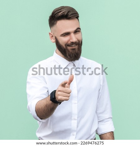 Hey, I choose you. Portrait of attractive flirting bearded businessman wearing white shirt pointing finger and winking at camera, choosing you. Indoor studio shot isolated on light green background.