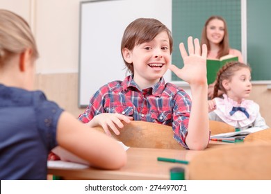 Hey Everyone. Happy Pupil Boy Waving With Hand Turned Away From Teacher In Classroom.