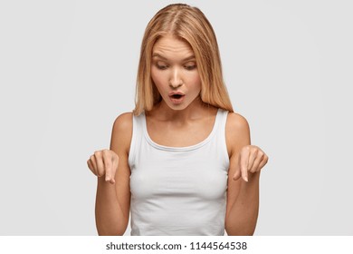 Hey, just look down! Pretty lovely young female with long straight hair, points on floor with fore fingers, notices something awful, wears casual white vest, stands alone in studio. People, reaction