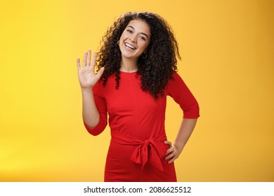 Hey My Name Is. Friendly-looking Self-assured Carefree Cute 25s Woman With Curly Hair Waving With Raised Palm In Hello Or Hi Gesture Smiling Broadly Greeting New Coworkers Over Yellow Background