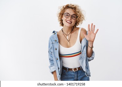 Hey nice to meet you. Portrait of friendly-looking sociable cute european female in glasses with short fair curly haircut waving raised palm and smiling cute as greeting and saying hi over white wall - Shutterstock ID 1401719258