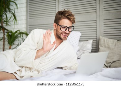 Hey there. Waist up portrait of smiling man communicating with friends and relatives online. He is lying in bathrobe on bed and waving at computer screen with hand - Powered by Shutterstock