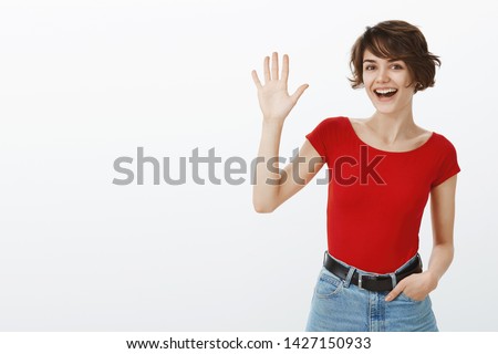 Hey whats up. Attractive sociable cheerful friendly european girl short haircut waving raised palm say hi hello greeting you welcoming guests stand white background meet new people