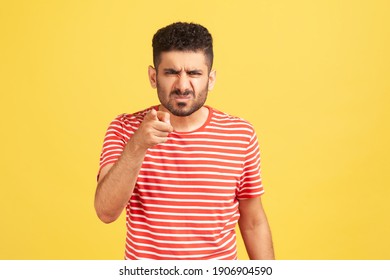 Hey you! Angry bossy man with beard in striped t-shirt grimacing pointing finger at camera, scolding and blaming you. Indoor studio shot isolated on yellow background